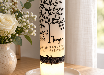 „Weiße Trauerkerze mit schwarzem Lebensbaum und LED-Licht auf einem Holztisch neben weißen Blumen.“