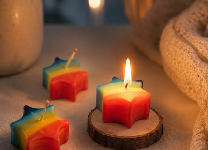 Angezündete Regenbogen-Sternkerze auf Holzteller in warmem Abendlicht als kleines Friedens- und Hoffnungslicht.