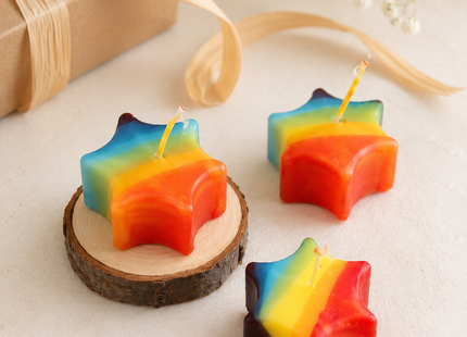 Drei Regenbogen-Sternkerzen auf hellem Holztisch, liebevoll dekoriert mit natürlichem Holzteller.